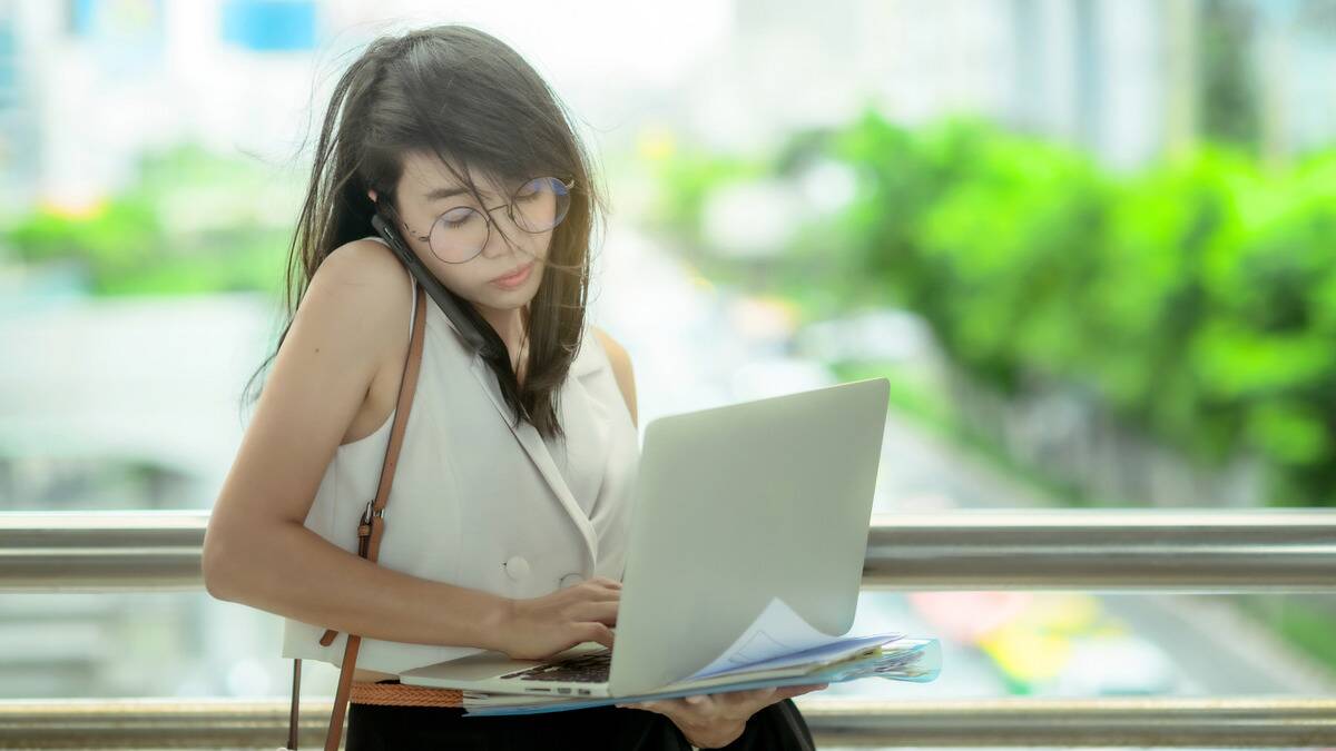 A woman balancing her open laptop on top of some messy notebooks, her hair touseled, shoulder holding her phone to her ear as she types and talks.