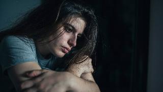 A close shot of a woman sitting and leaning forward with her arms folded, looking ahead with an empty expression.