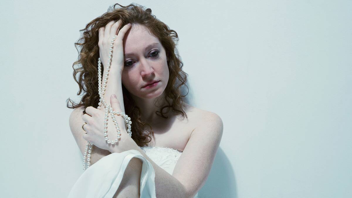 A woman sitting on the floor against a bare wall, knee drawn up one hand on her forehead as it holds a long pearl necklace. She's looking toward the floor with a tired expression.