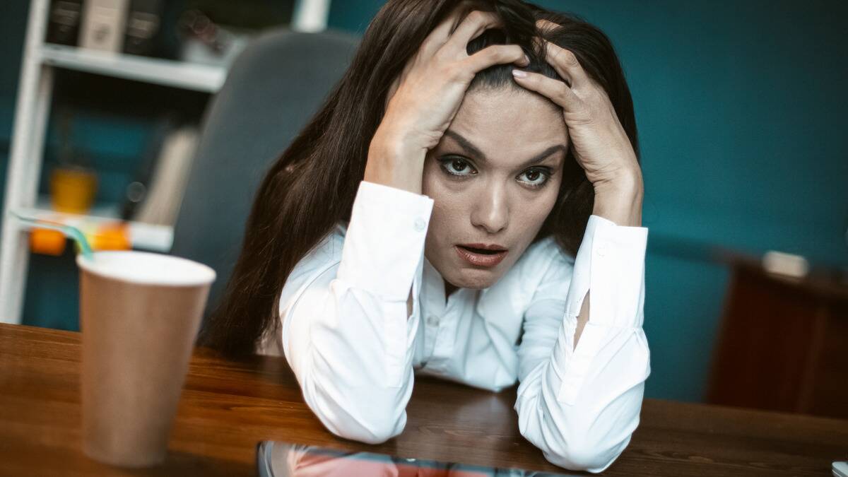 A close, tilted shot of a woman leaning forward, elbows on her desk, hands in her hair, looking very stressed.