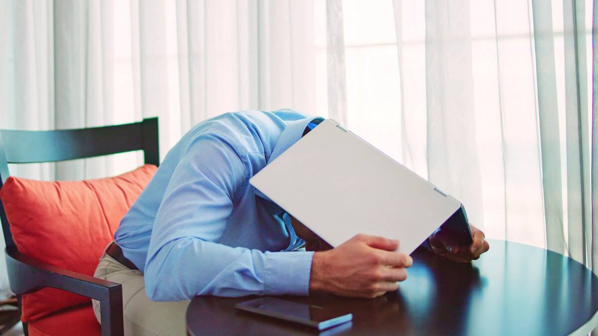 A man leaning forward on his desk, laptop covering his head.