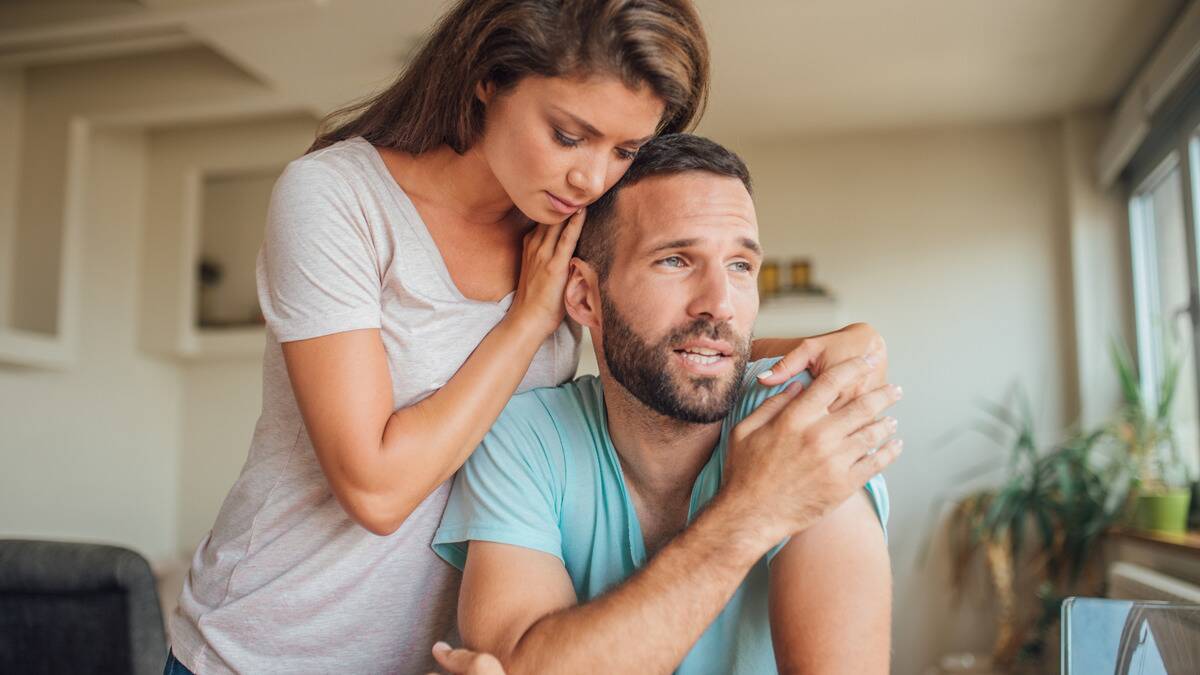 A man looking forward sadly as he sits, his girlfriend standing above him and embracing him from behind.