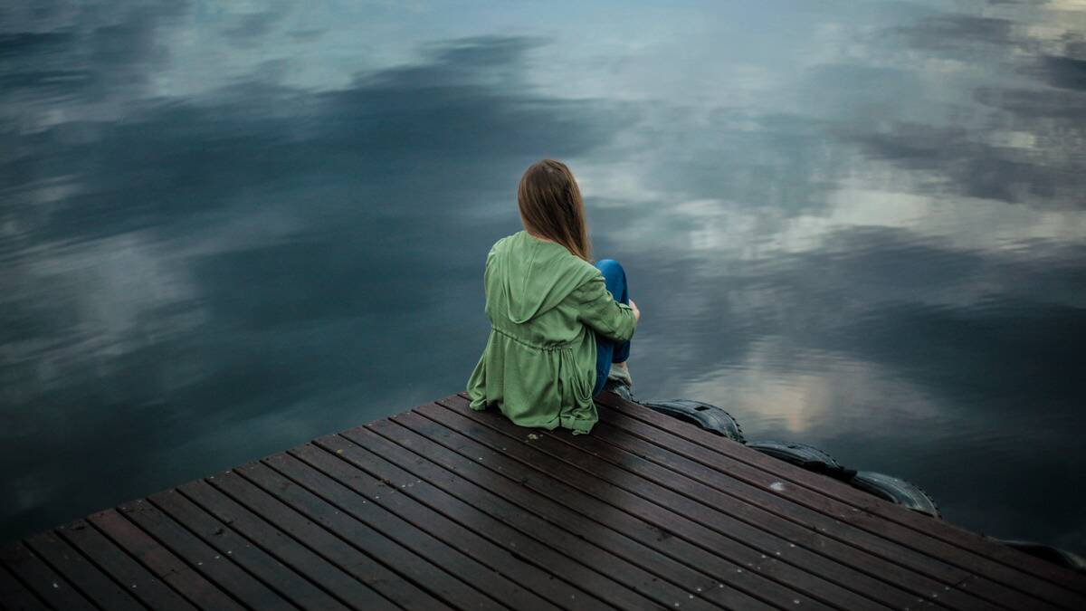 A woman sitting on the corner of a small dock in front of still grey-blue water.