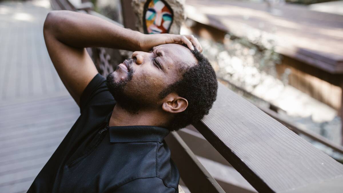 A man sitting on a bench outside, head tilted back, a hand up on his forehead, eyes closed in frustration.