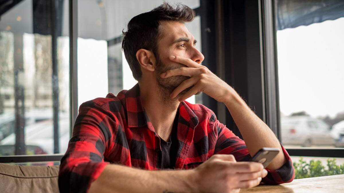 A man sitting at a table, looking out the window with a pained expression, one hand over his mouth, the other holding his phone.