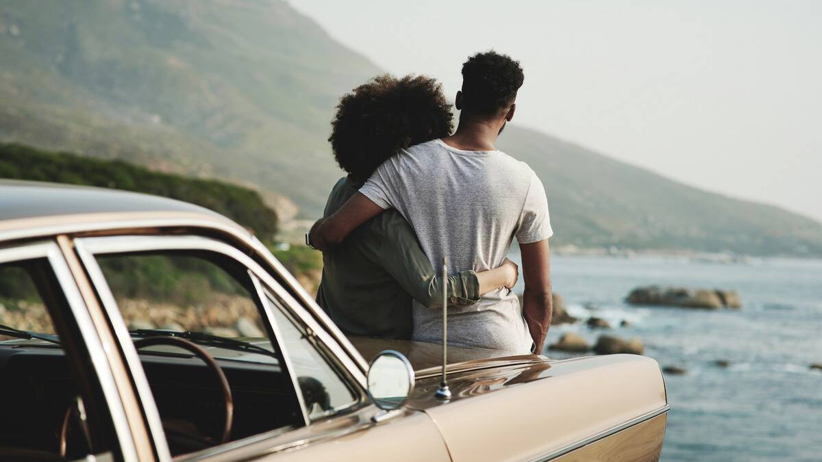 A couple photographed from behind as they stand, arms around each other, looking out at the scenic horizon, leaning against the hood of their car.