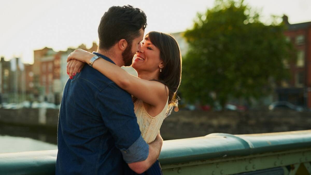 A couple standing outside on a bridge, arms around each other as they go in for a kiss.