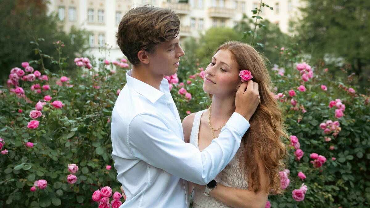 A young couple standing outside, the man putting a pink flower in the woman's hair, tucked behind her ear.