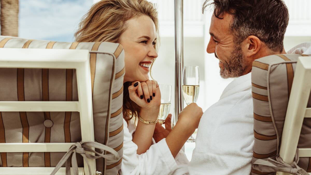 A close shot from behind of a couple sitting in beach chairs next to one another, each holding a glass of champagne, leaning toward one another to talk.