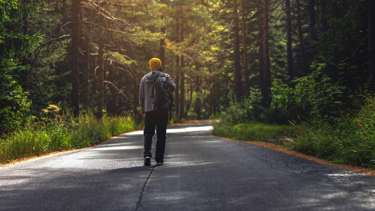 A man walking down a tree-lined road with beams of light shining through, facing away from the camera, a bag hiked over one shoulder.