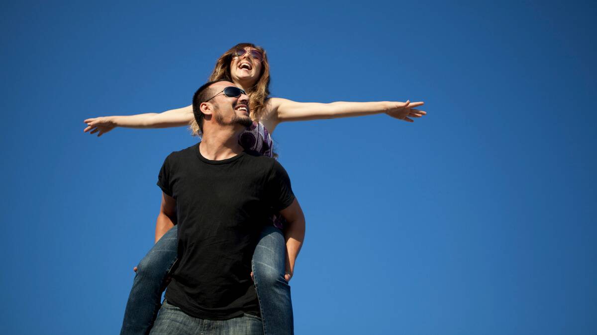 A man giving his girlfriend a piggyback ride under a bright blue sky. Her arms are out, both are smiling and laughing.