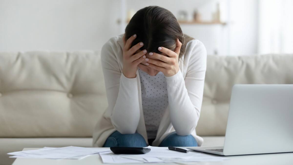 A woman sitting at her coffee table that's covered in papers, a calculator, her laptop, and a pen, leaning over with her head in her hands.