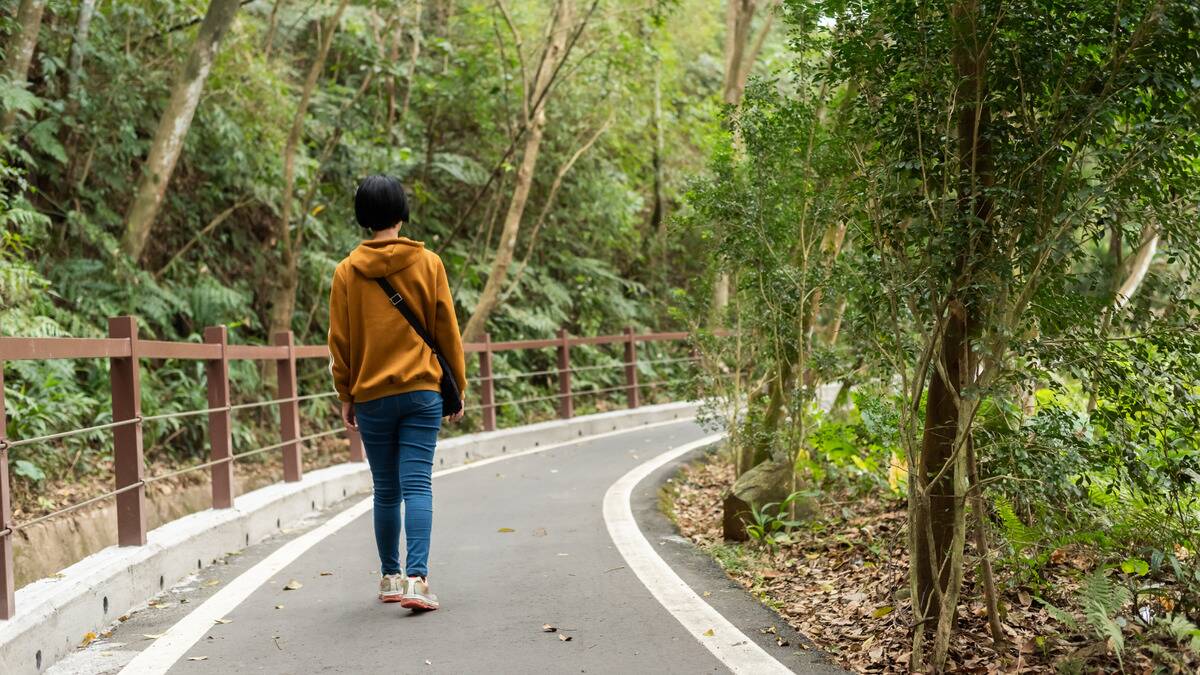 A woman walking away from the camera down a paved walking path surrounded by trees. 