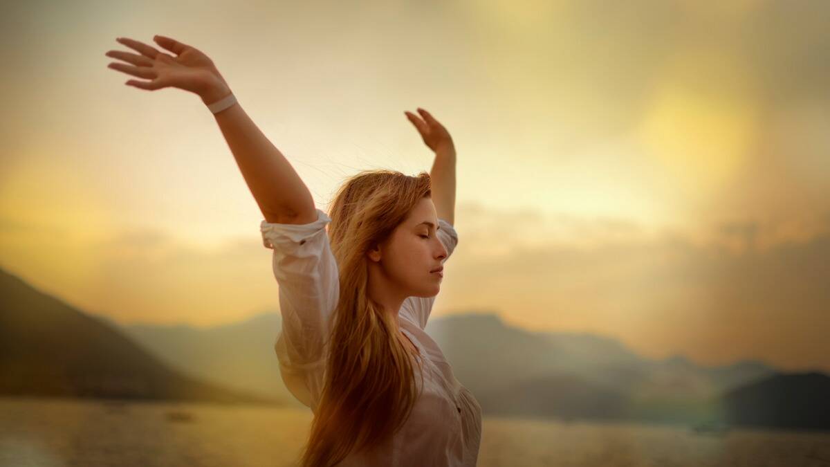 A woman standing outside with her eyes closed, arms up in the air.