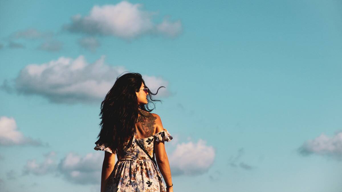 A woman standing beneath a bright blue sky, facing away from the camera but looking over her shoulder so we see her face in profile.