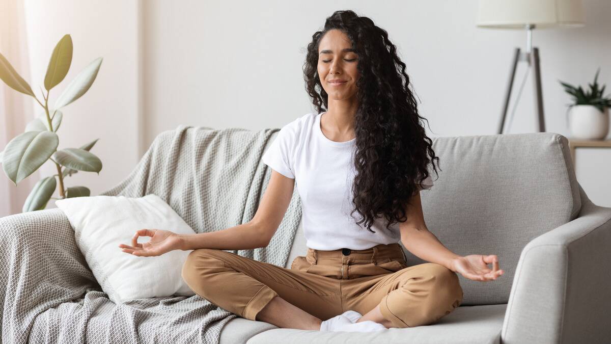 A woman sitting on her couch meditating, legs crossed, arms poised on her knees, eyes closed with a gentle smile.