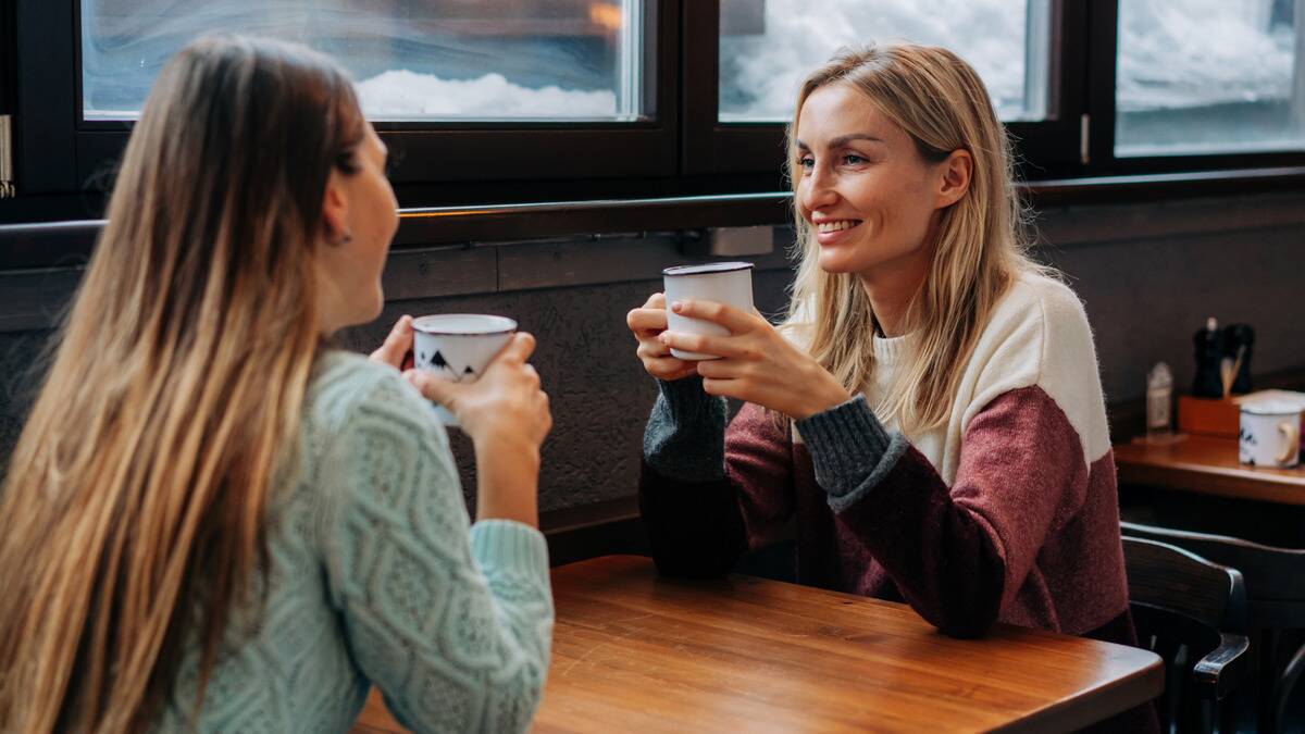 Two women sat across from each other at a cafe, each holding a mug of coffee.