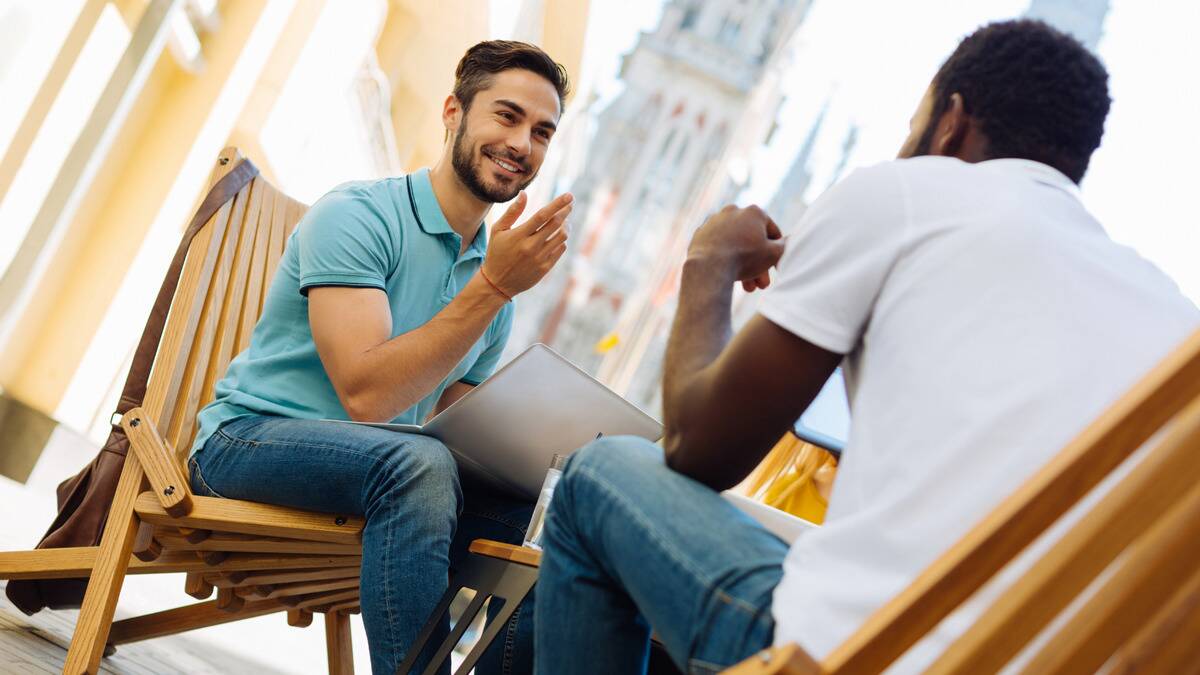 A low-angle shot of two men sitting opposite one another at a bistro or cafe, gesturing with their hands as they chat.