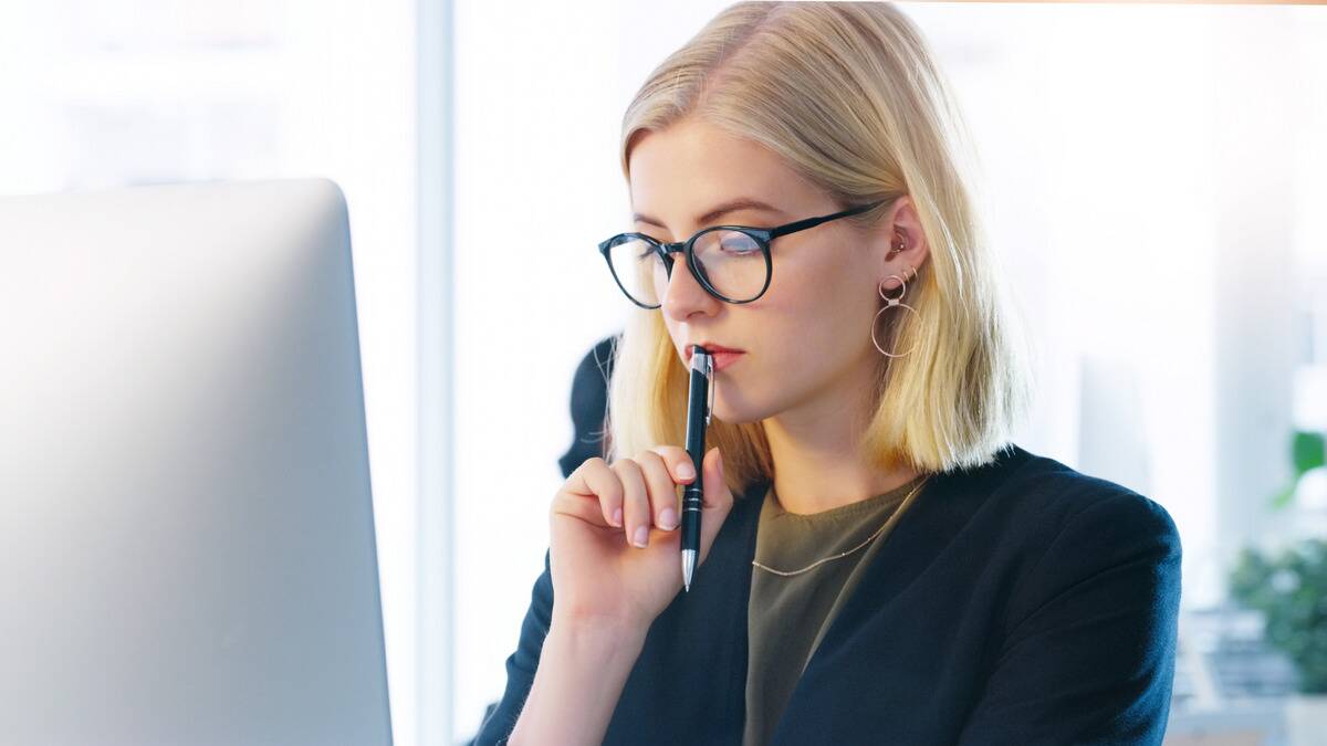 A close shot of a business woman sitting at her desk, her pen pressed against her lips, looking down slightly, clearly thinking something over.