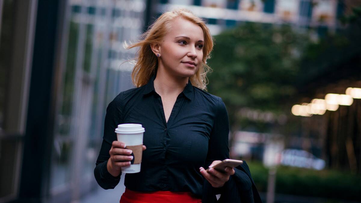 A woman holding a coffee and her cell phone, looking off to the side as she walks down a city street.