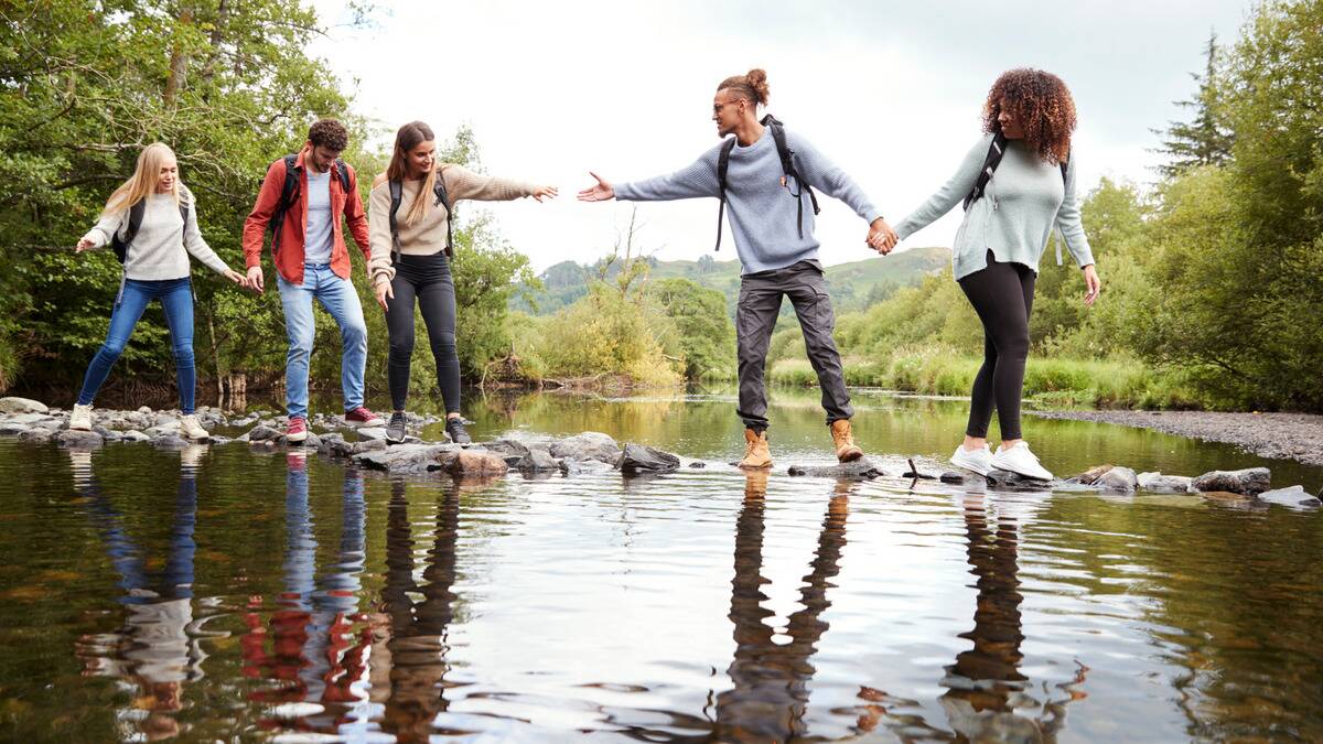 A group of friends crossing a creek while on a hike by walking across some rocks, one reaching out across the rocks to give a hand to another.