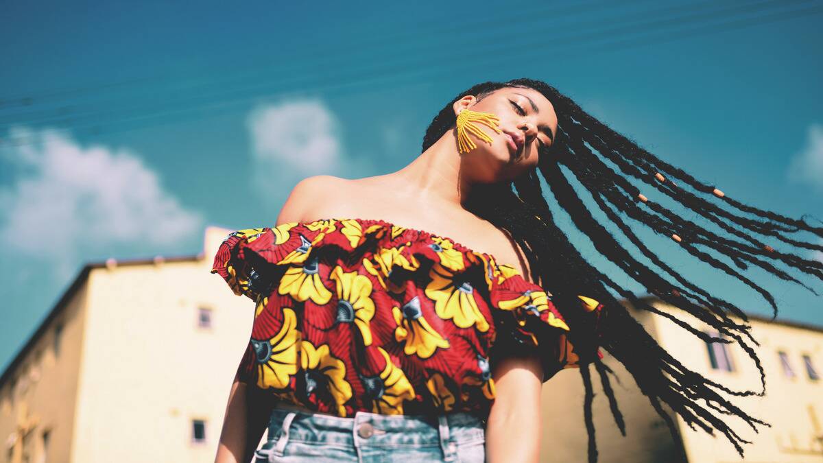 A low-angle photo of a woman with her eyes closed swinging her long braids out to the side under a bright blue sky.