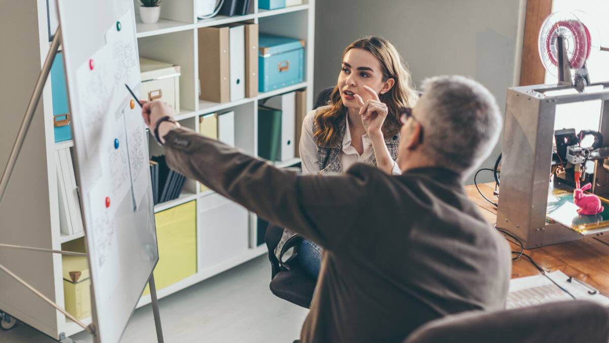 An employee and her boss in a meeting, the boss pointing something out on a board while the employee speaks.