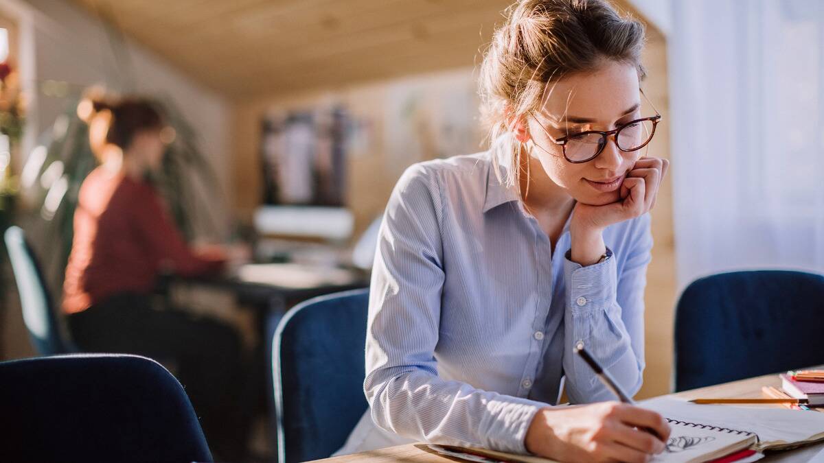 A woman sitting at her desk at work, smiling as she doodles in a sketchbook.