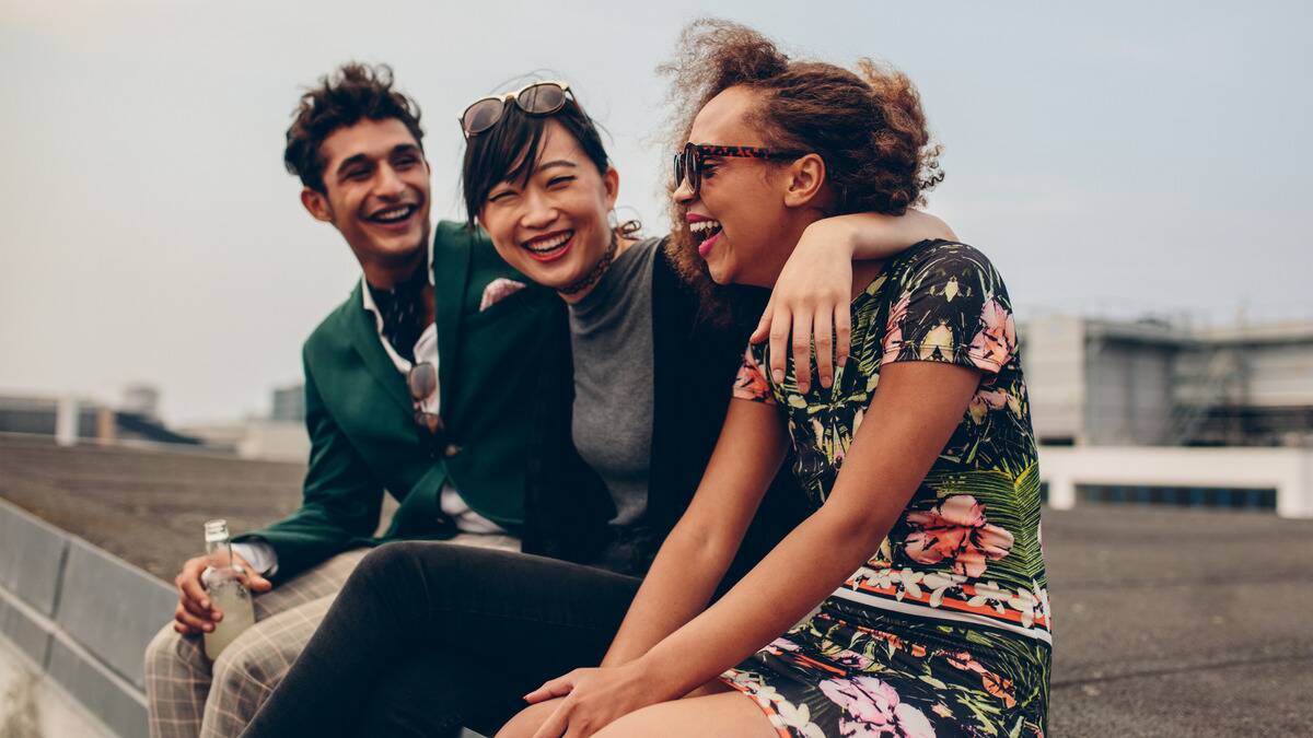 A trio of friends outside, sitting on a rooftop, arms around one another, all smiling.