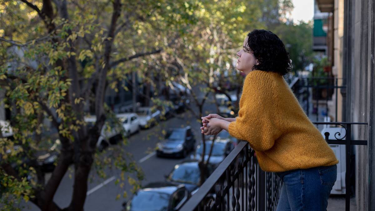 A woman standing on her balcony looking out to the city street and trees planted along it.