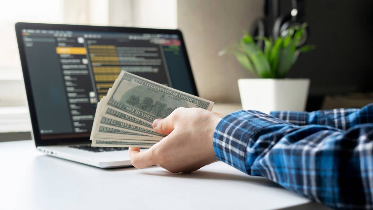 Someone holding a stack of American bills in front of their open computer.