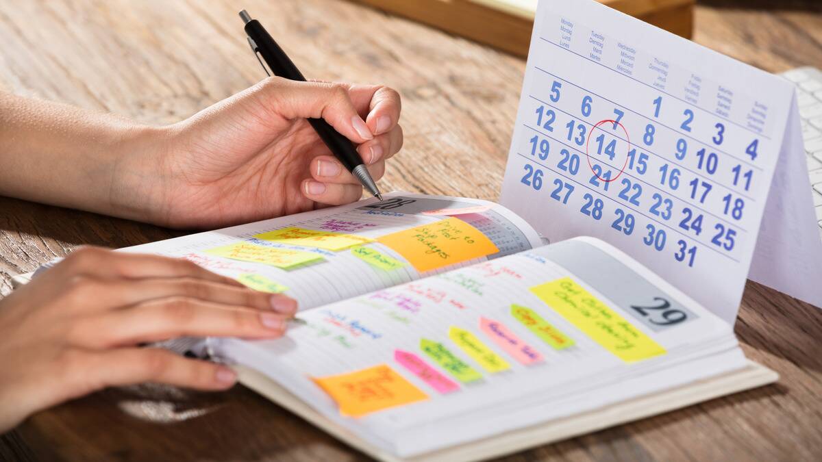 Someone writing a schedule in a journal while cross referencing a monthly desk calendar.