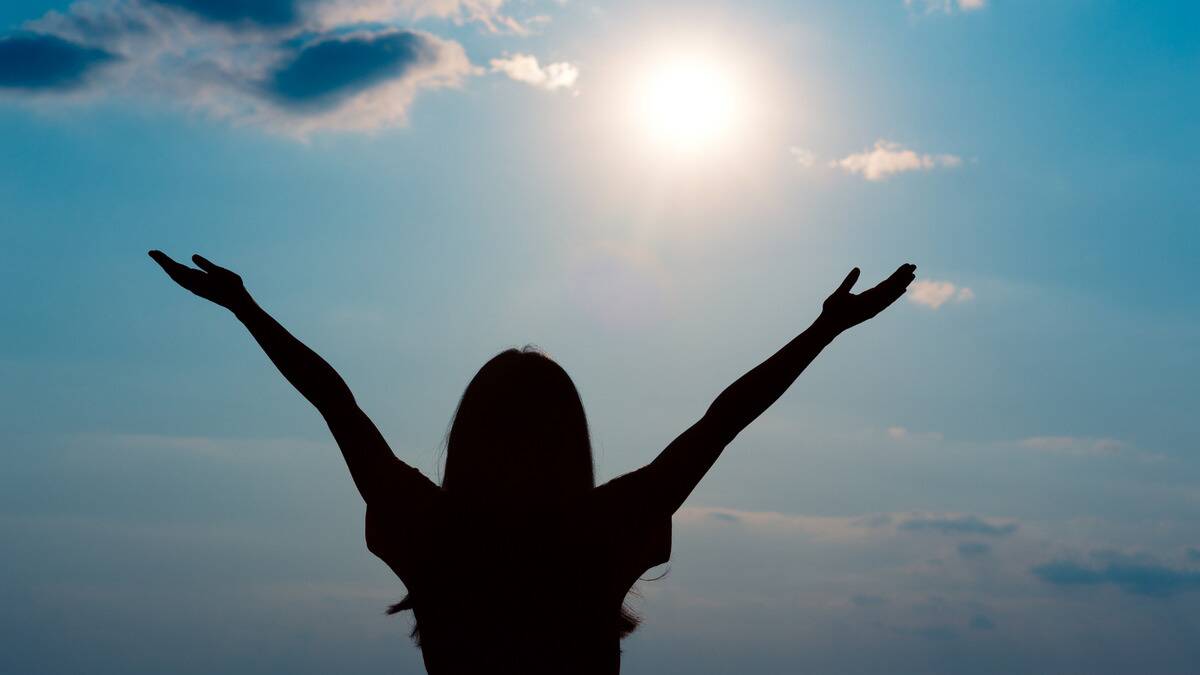 The silhouette of a woman standing with her arms out as she looks to the sunny sky.