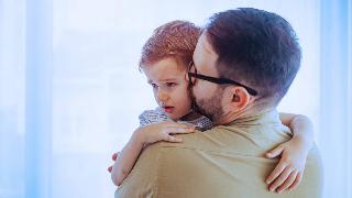 A father holding and hugging his young son, who looks distressed, kissing his cheek.