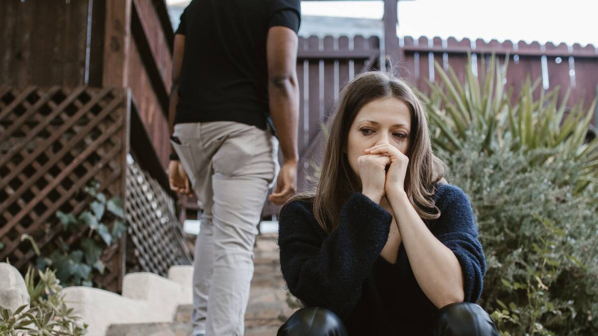 A woman sitting on some steps outside, a man seen walking away from her in the background.