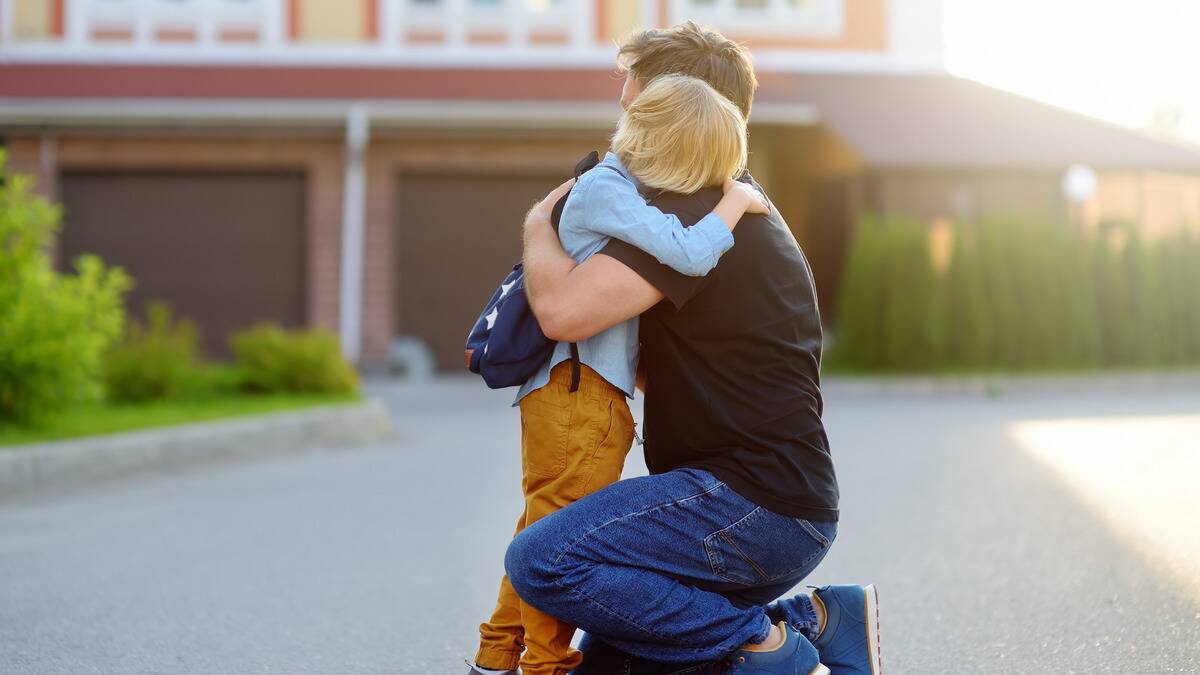 A father kneeling down to hug his young son outside their home, presumably before he goes off to school.