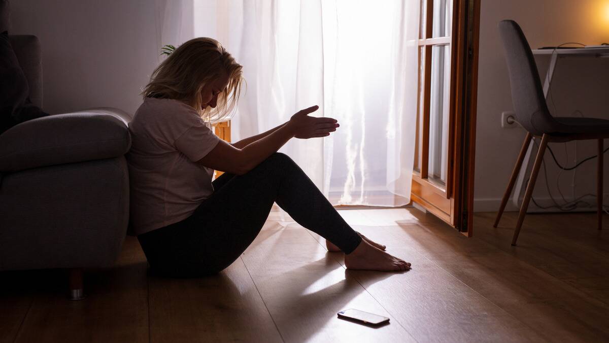 A woman sitting on the floor, her phone on the ground, knees drawn up, hands together, looking tired and fed up.