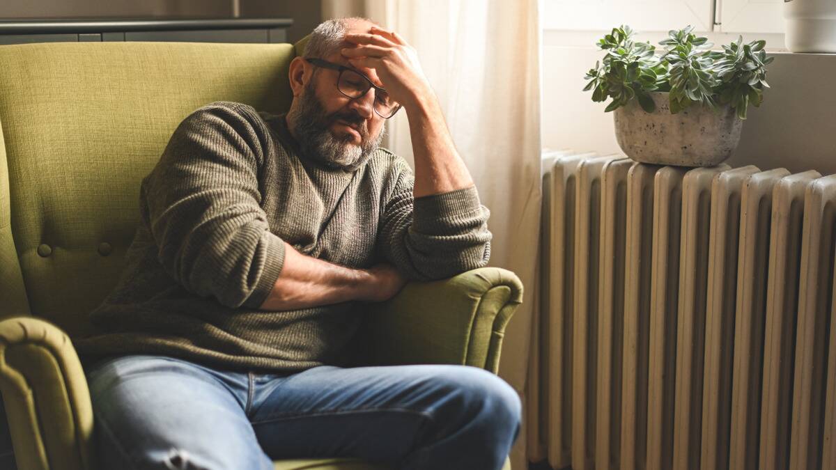 A man sitting on a chair in his home, hand on his forehead, looking frustrated.