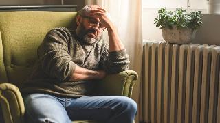 A man sitting on a chair in his home, hand on his forehead, looking frustrated.