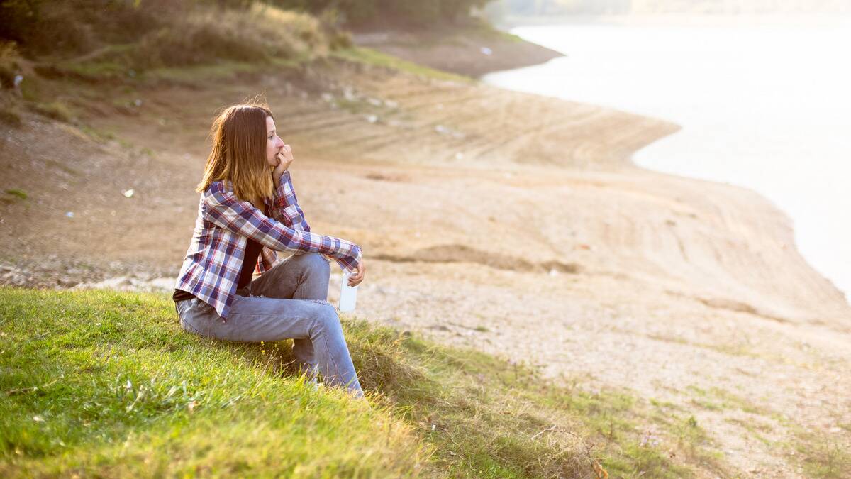 A woman sitting out by the beach, a hand on her chin, looking forward pensively.