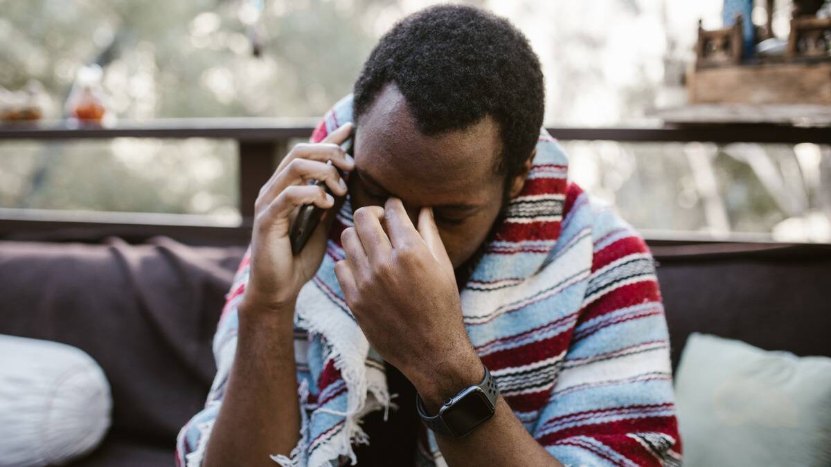 A man sitting outside on the phone, head forward, hand pinching the bridge of his nose, a blanket around his shoulders.