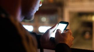 An over-the-shoulder shot of a man texting on his phone, the bright screen illuminating his hands and face.