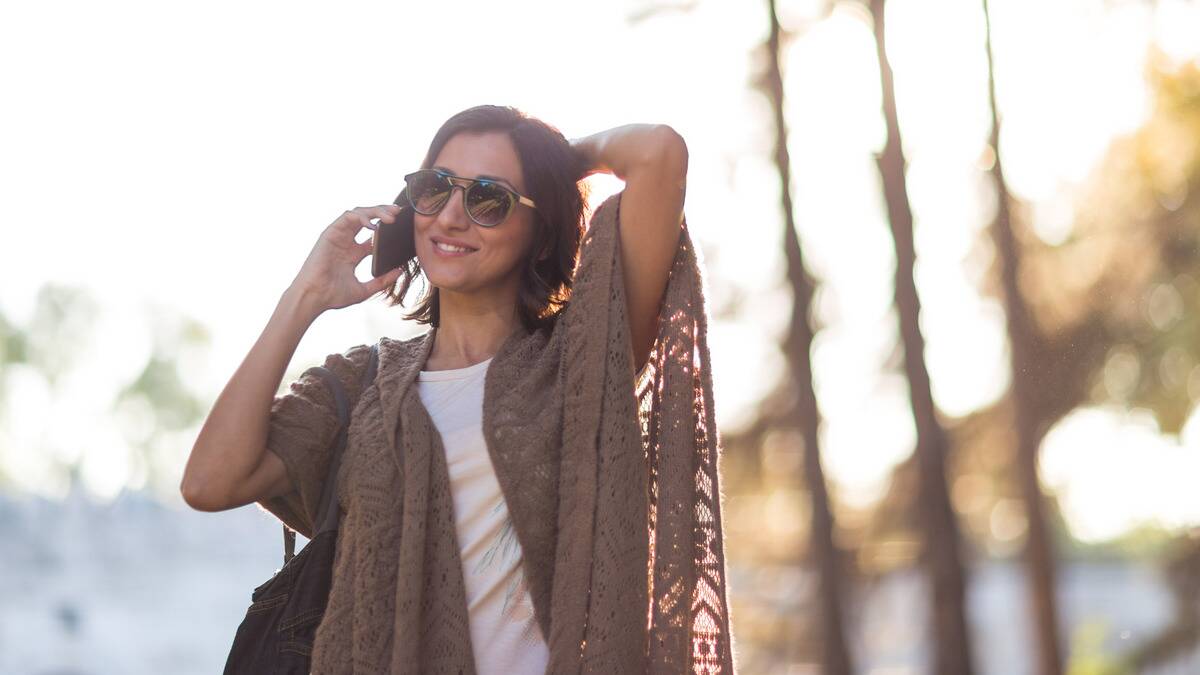 A woman speaking on the phone as she walks outside, her free hand in her hair, a smile on her face.