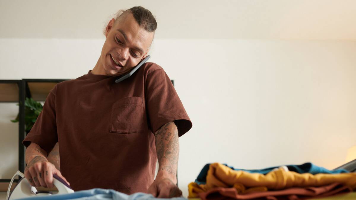 A man talking on the phone while ironing some clothes, holding the phone between his cheek and his shoulder.