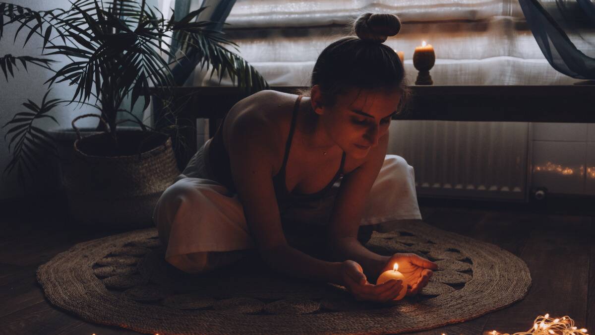 A woman sitting cross legged on the floor, leaning forward, cupping her hands around a lit candle.