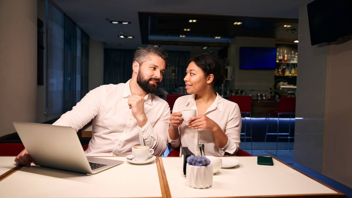 Two coworkers, one man and one woman, sitting side by side at a diner table. The man has his laptop open and is smiling as he leans toward his coworker, who's also smiling and holding a mug.