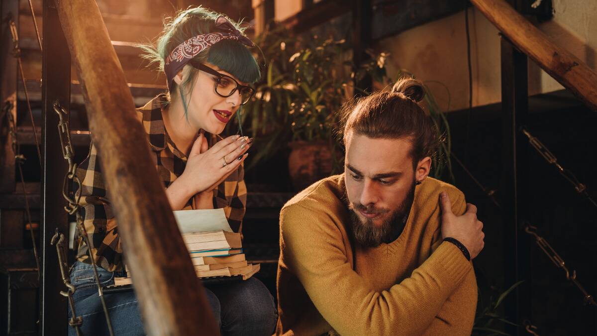 A man and a woman sitting on a set of indoor stairs, the woman with her hands together making a pleading expression toward the man, who's looking downward as he thinks.