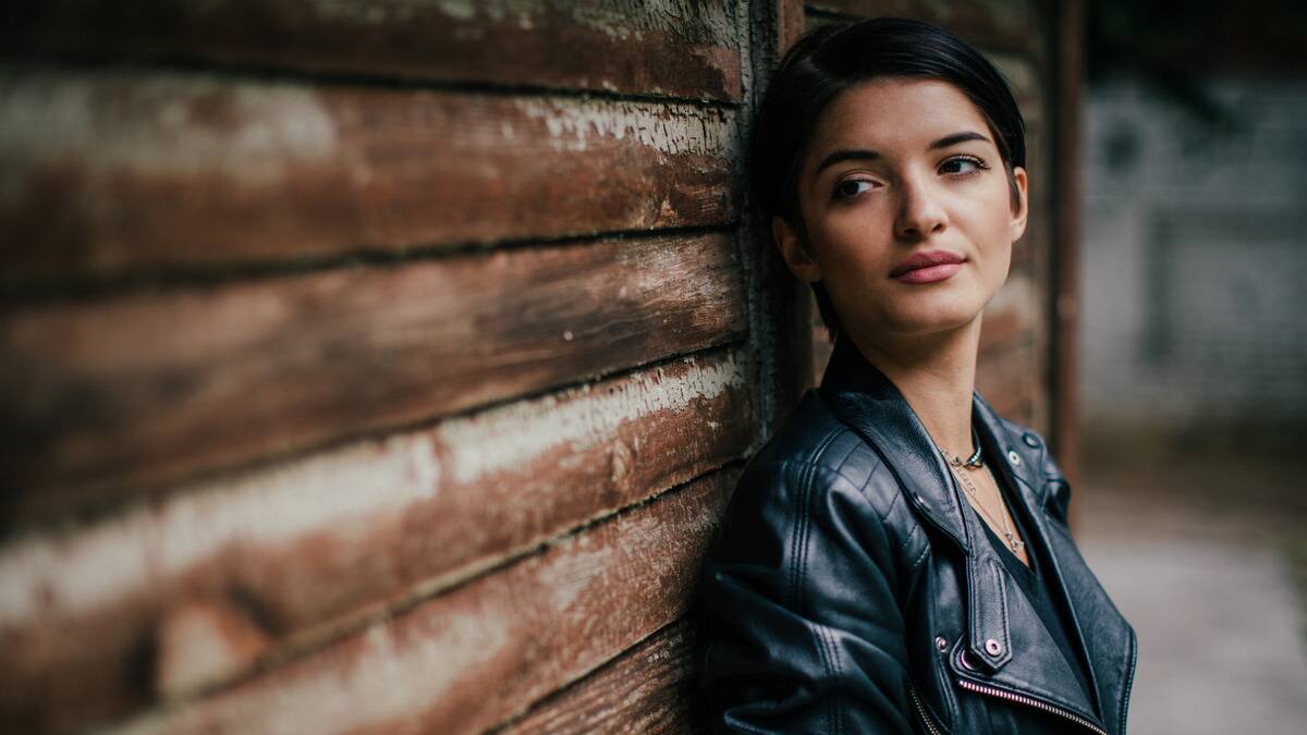 A woman standing outdoors, leaning against a wall, looking off to the side coolly.