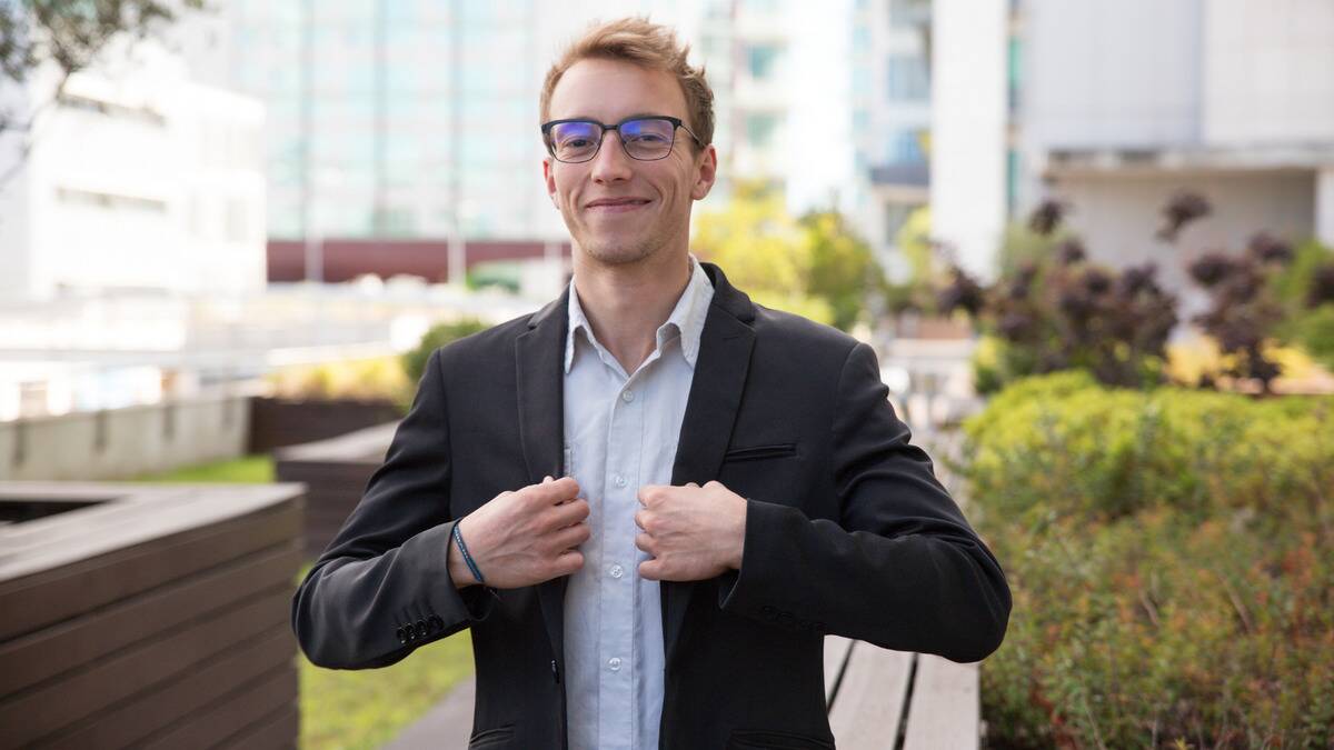 A man walking outside, smiling confidently, his hands on his blazer collar.