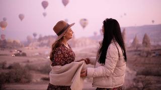 Two women standing in front of a field, holding both pairs of hands together, smiling, watching hot air balloons rise in the background.